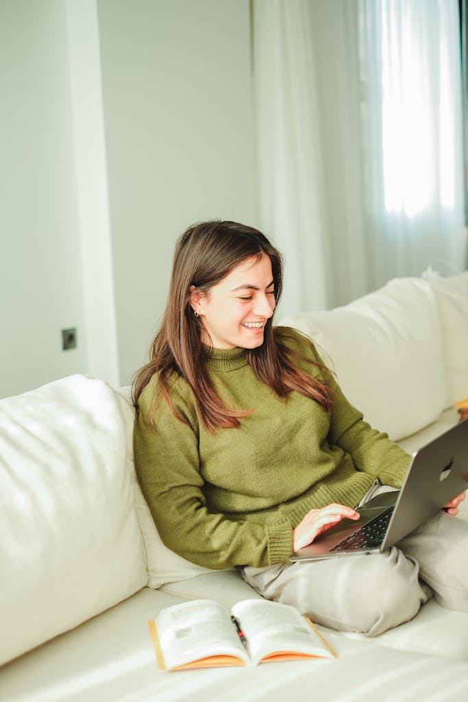 Photo by Gülşah Aydoğan A young woman enjoys working on her laptop while relaxing on a sofa indoors, surrounded by books.