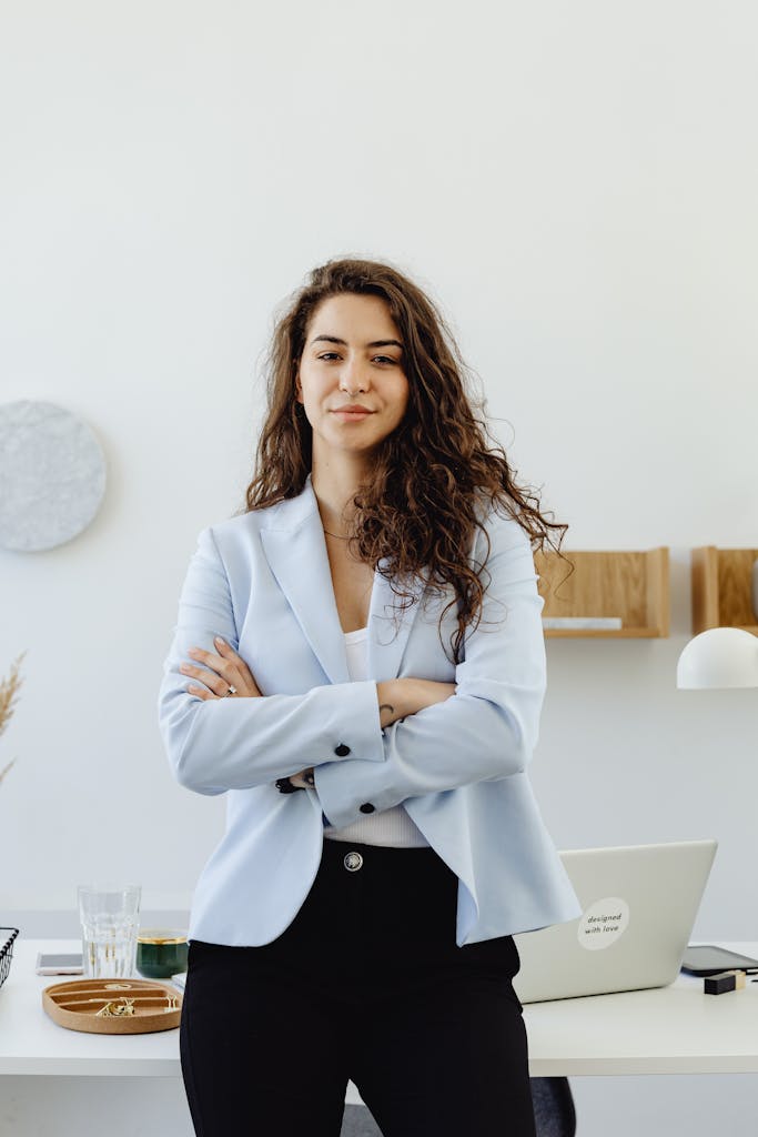 Photo by Photo By: Kaboompics.com Professional woman in a white blazer standing confidently in an office with arms crossed.