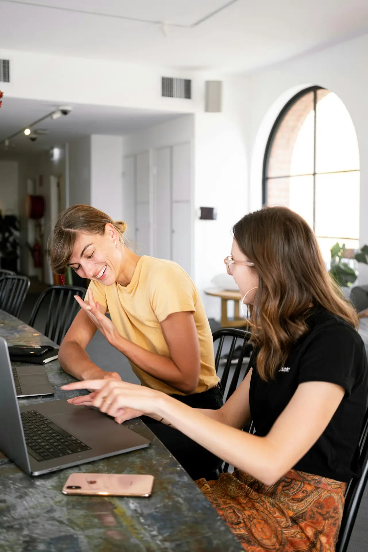 Photo by Canva Studio Two women engaged in a collaborative discussion at a modern office setting over laptops.